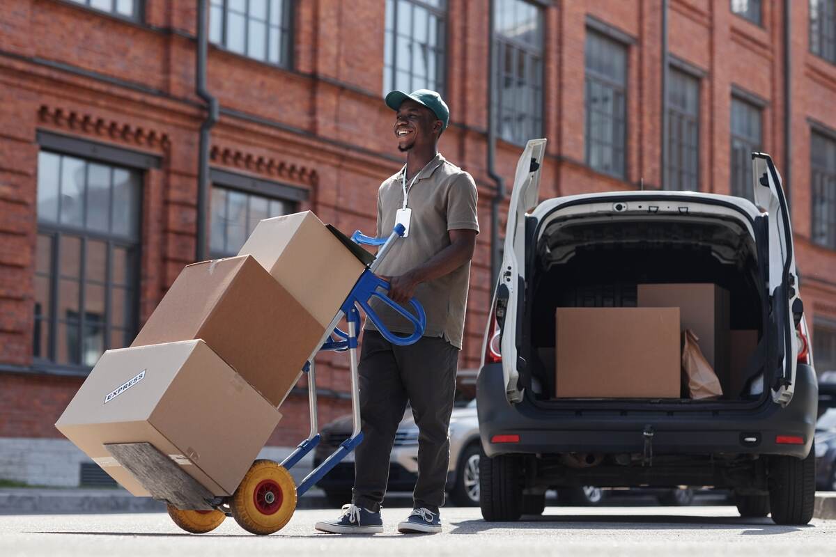 Smiling delivery worker moves boxes on a hand truck near a parcel van.
